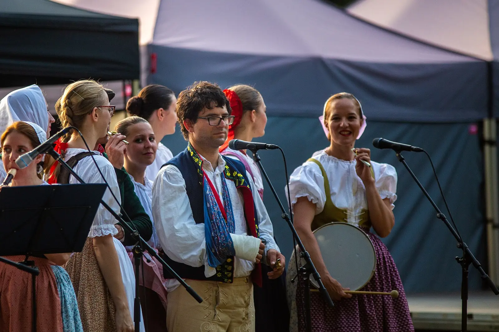 Members of a folk ensemble in Moravian costume on a festival stage, singing into microphones, one holding a small frame drum
