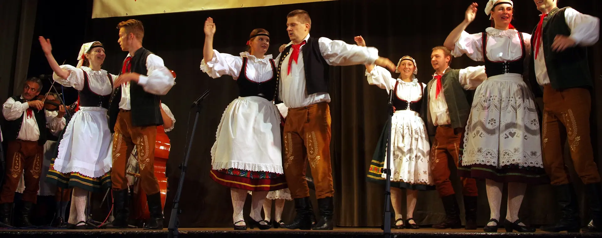 Adult folk dancers on a stage in Czech costume, women in white aprons and black bodices and men in dark waistcoats with red neckerchiefs, arms raised in a dance, with a violinist and a double bass at the left
